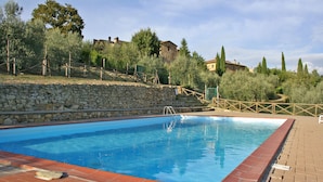 Una piscina rettangolare con un muro in pietra e una recinzione in legno su un lato, circondata da alberi e edifici sullo sfondo.