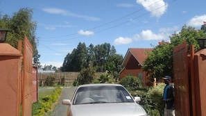 A residential street with a parked car, a person standing by a gate, and houses with red roofs. - Watercrest Gardens (Eldoret)