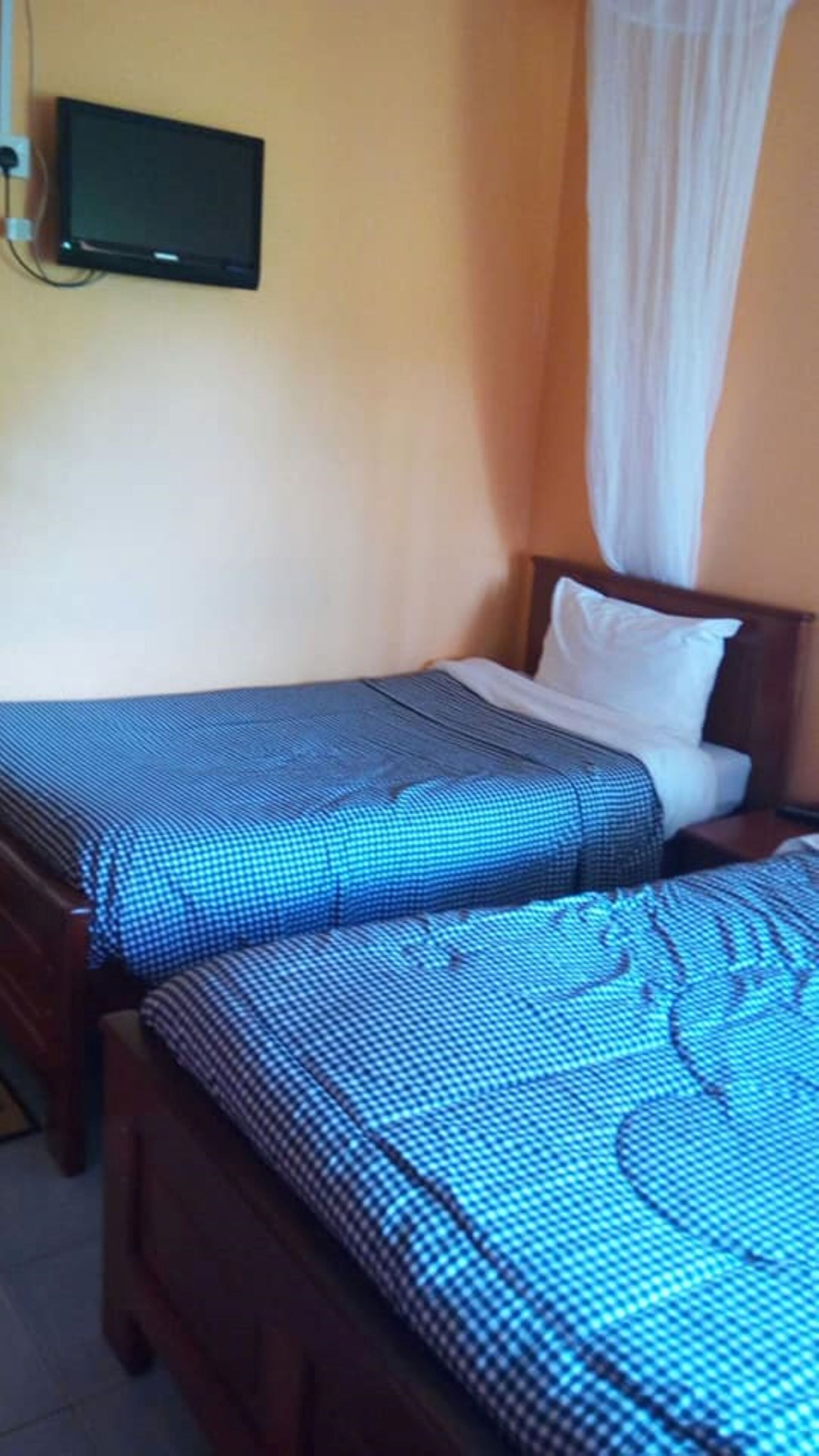 A bedroom with two single beds, a television mounted on the wall, and a white curtain.