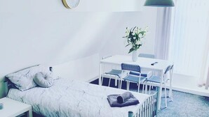 Ein kleiner, ordentlicher Raum mit einem Bett, einem Schreibtisch und einem Stuhl. Es gibt ein Fenster mit Vorhängen, eine Pendelleuchte und eine Uhr an der Wand.