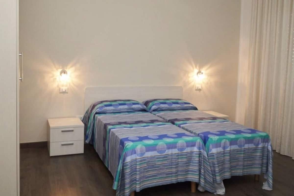 A room with two beds, each with a striped bedspread, a bedside table with drawers, and two wall-mounted lights.