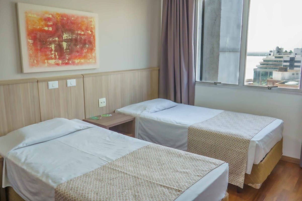 A hotel room with two single beds, a small table, a wall-mounted light switch, and a window with curtains.