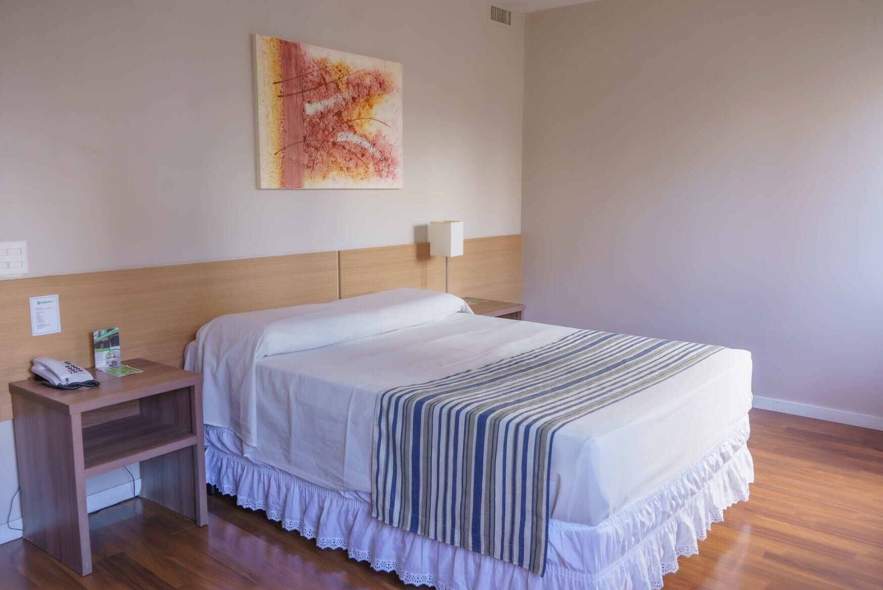 Een hotelkamer met een bed, nachtkastje, telefoon, lamp en een schilderij aan de muur.