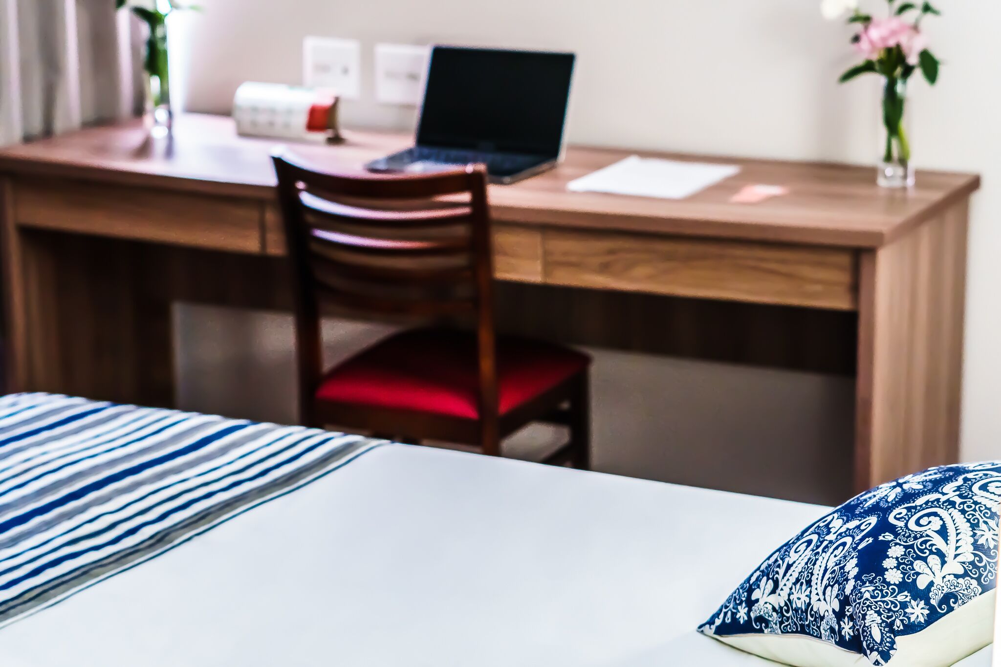 A hotel room with a wooden desk, a laptop, a chair with a red cushion, and a bed with striped bedding.