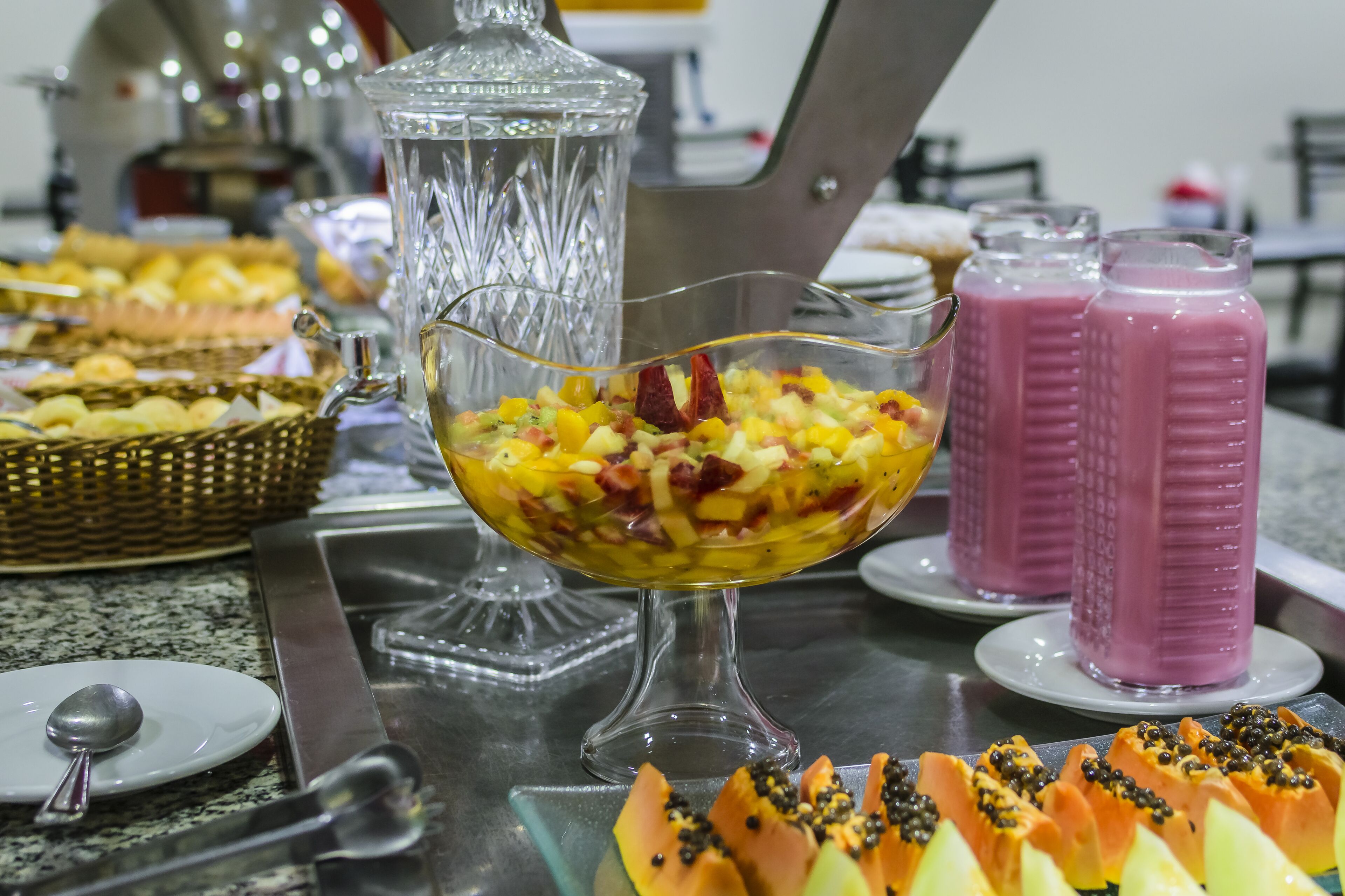 Un buffet con diversi cibi, tra cui una ciotola di insalata di frutta, un frullatore e un piatto con frutta a fettine.