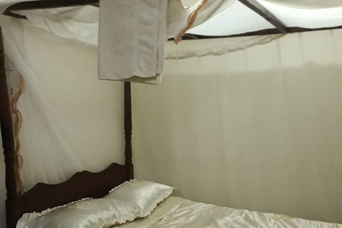 Una cama individual con edredón blanco y dos almohadas, cabecera de madera y dosel con tela blanca.