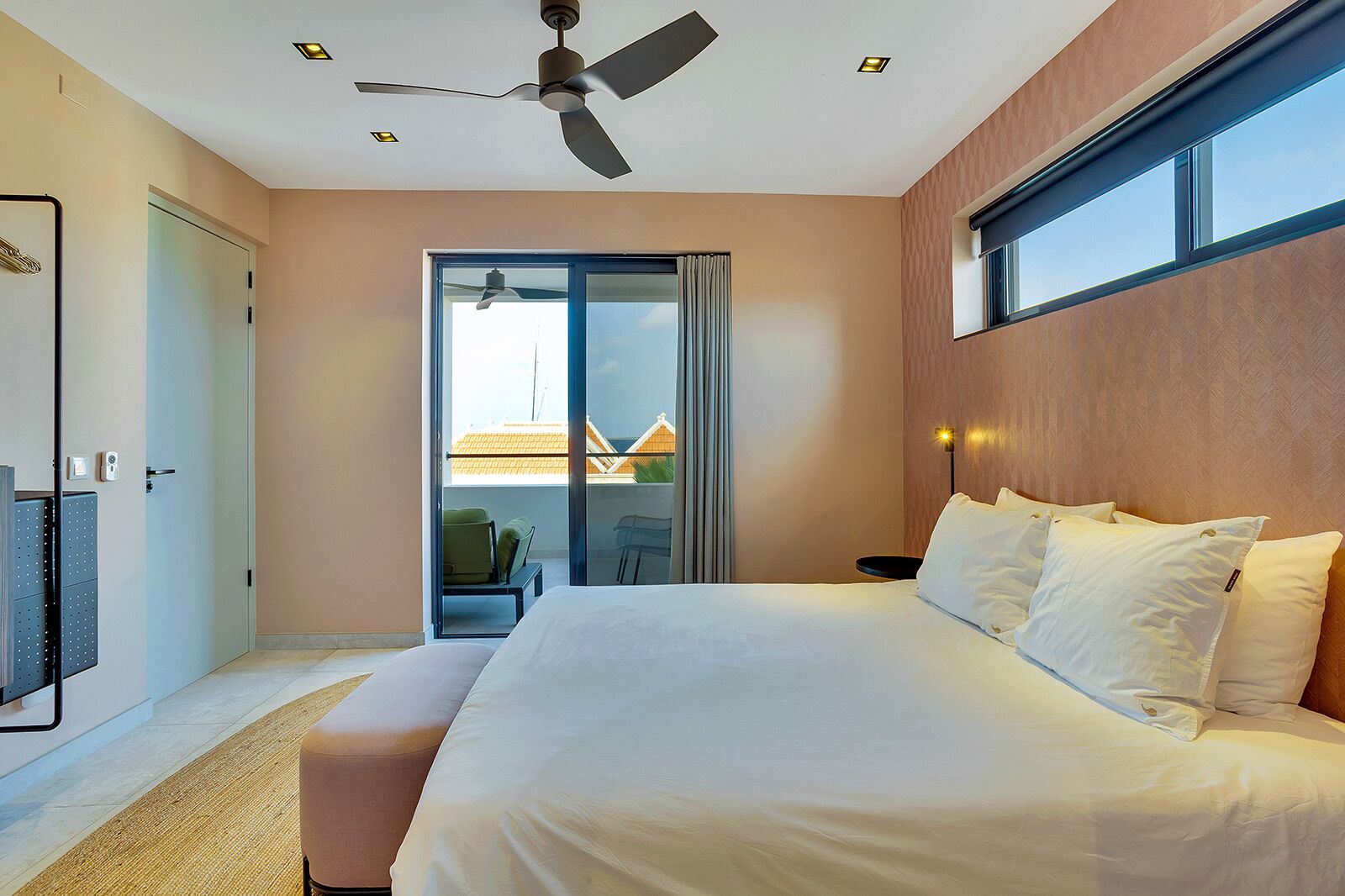Habitación de hotel con una cama grande, ventilador de techo, puerta corrediza de vidrio a un balcón y vistas de edificios.