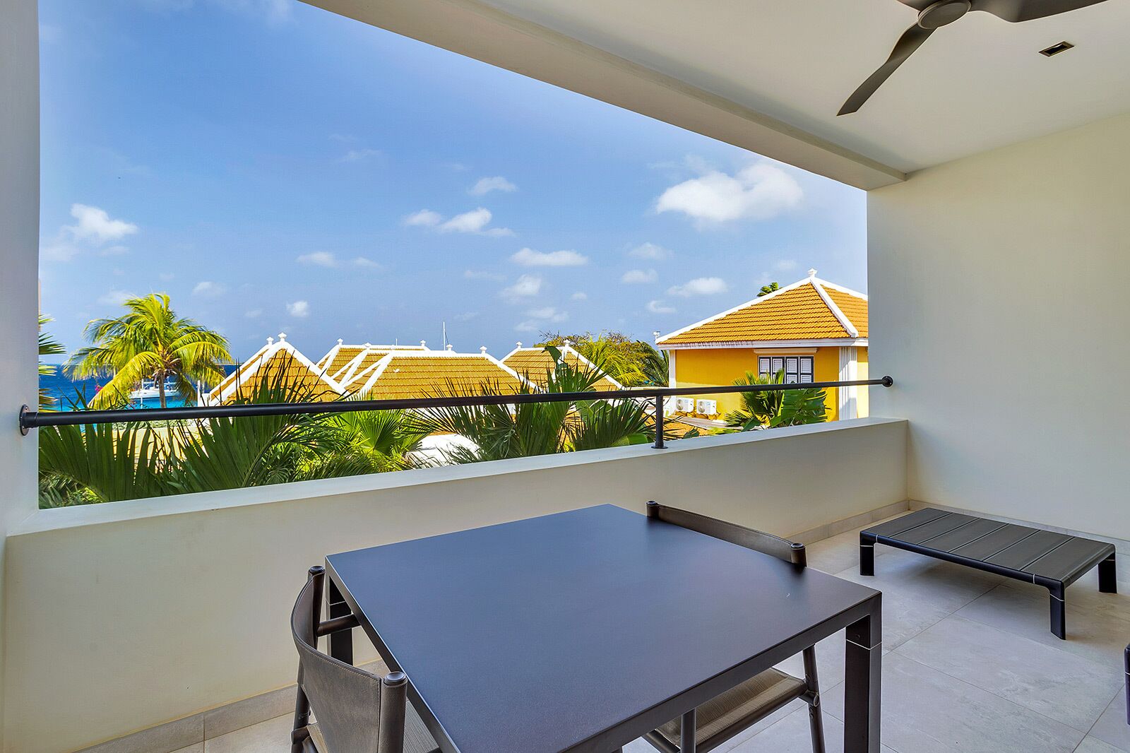 A balcony with a view of a yellow house, palm trees, and a clear blue sky.
