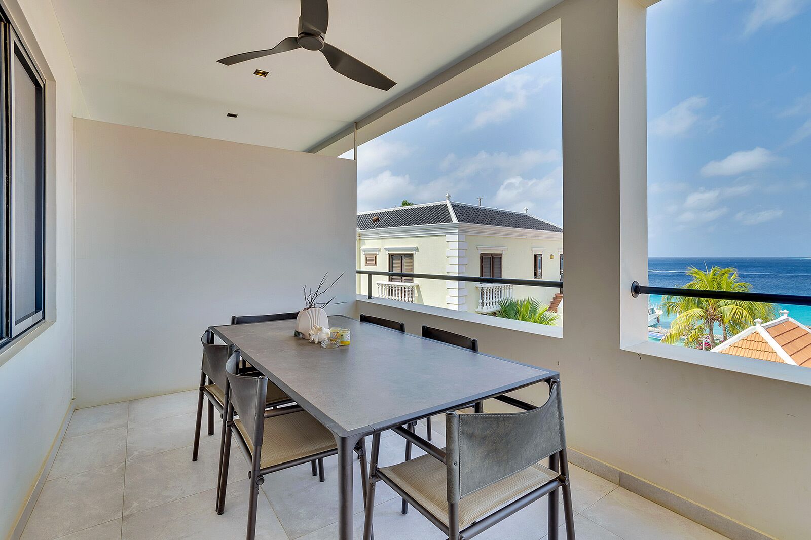 Un balcon avec une table et des chaises pour prendre les repas, un ventilateur de plafond et une vue sur un immeuble et l’océan.