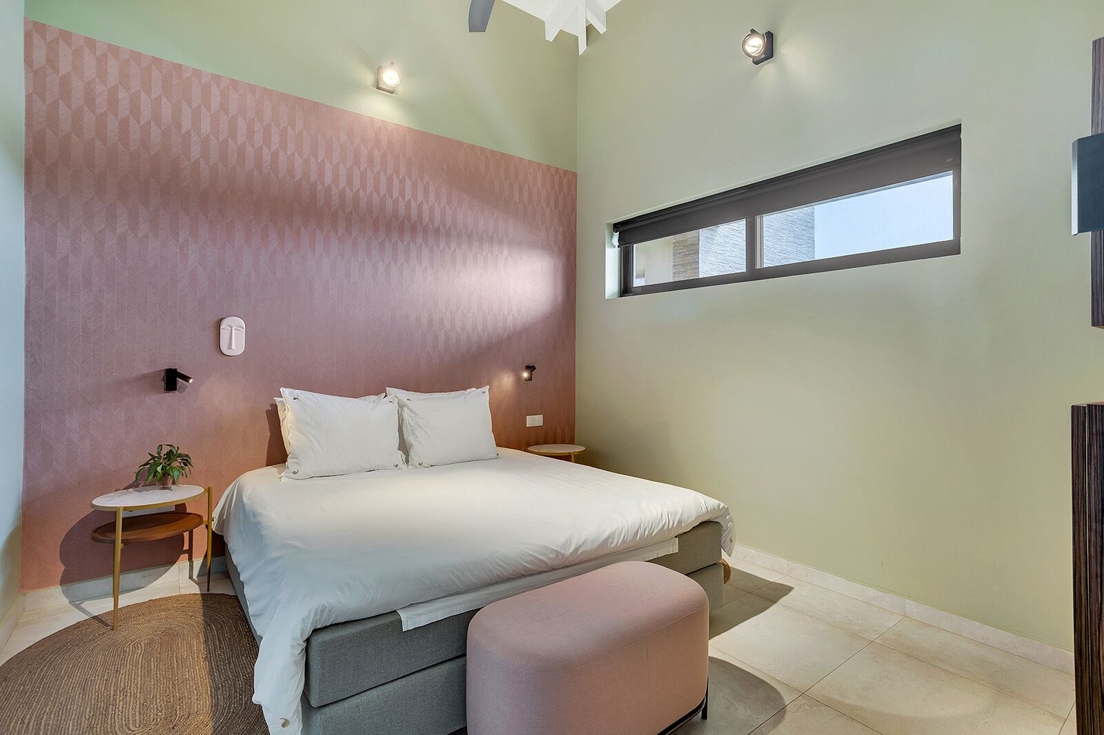 A bedroom with a bed, bedside tables, a small table with a plant, a window, and a wall-mounted light fixture.