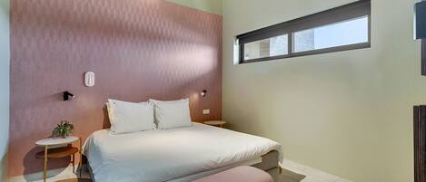 A bedroom with a bed, bedside tables, a small table with a plant, a window, and a wall-mounted light fixture.