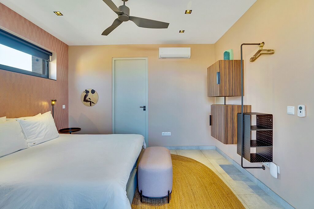 A hotel room with a bed, a standing fan, a small table, a wall-mounted shelf, and a door.