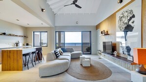 A modern living room with a large sofa, a dining area with bar stools, a kitchen with a wooden island, and a large window offering a view of the sea.