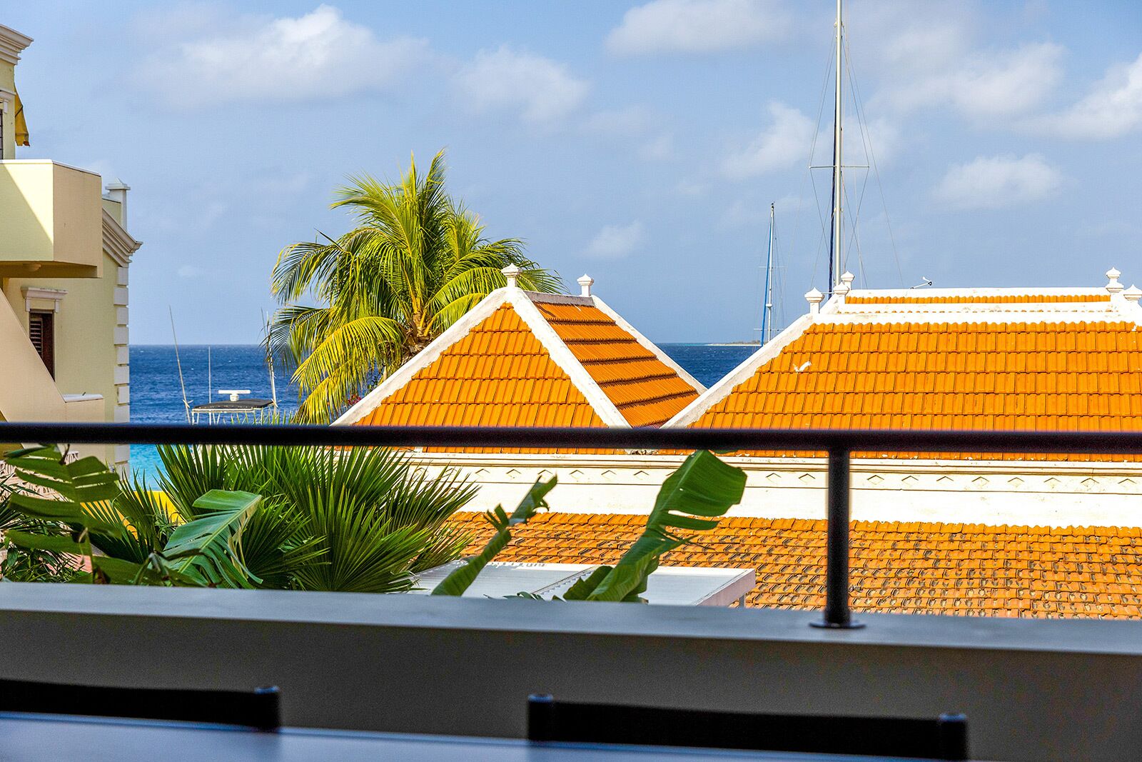 A coastal building with orange tiled roofs, a balcony with plants, and a view of the sea.