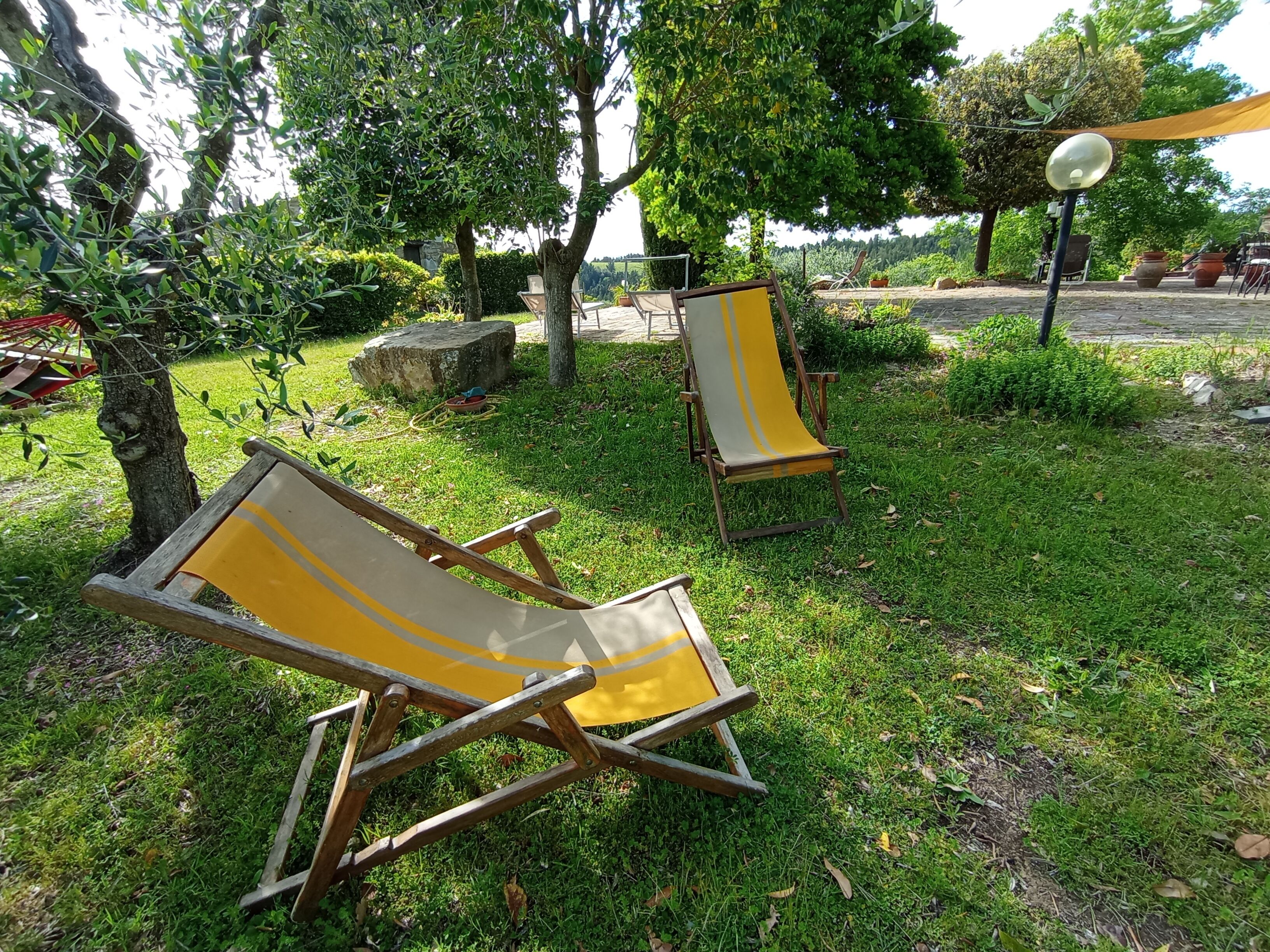Zwei hölzerne Liegestühle mit gelben Kissen in einer grünen Umgebung mit Bäumen und einem Steinweg.
