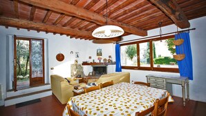 Une pièce chaleureuse avec un plafond en bois, une table à manger recouverte d’une nappe à motifs floraux, un canapé, une cheminée et une vue sur l’extérieur grâce à de grandes fenêtres.