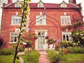 Une maison en briques rouges avec une porte verte, des chaises blanches devant et un chemin de jardin menant à l’entrée.