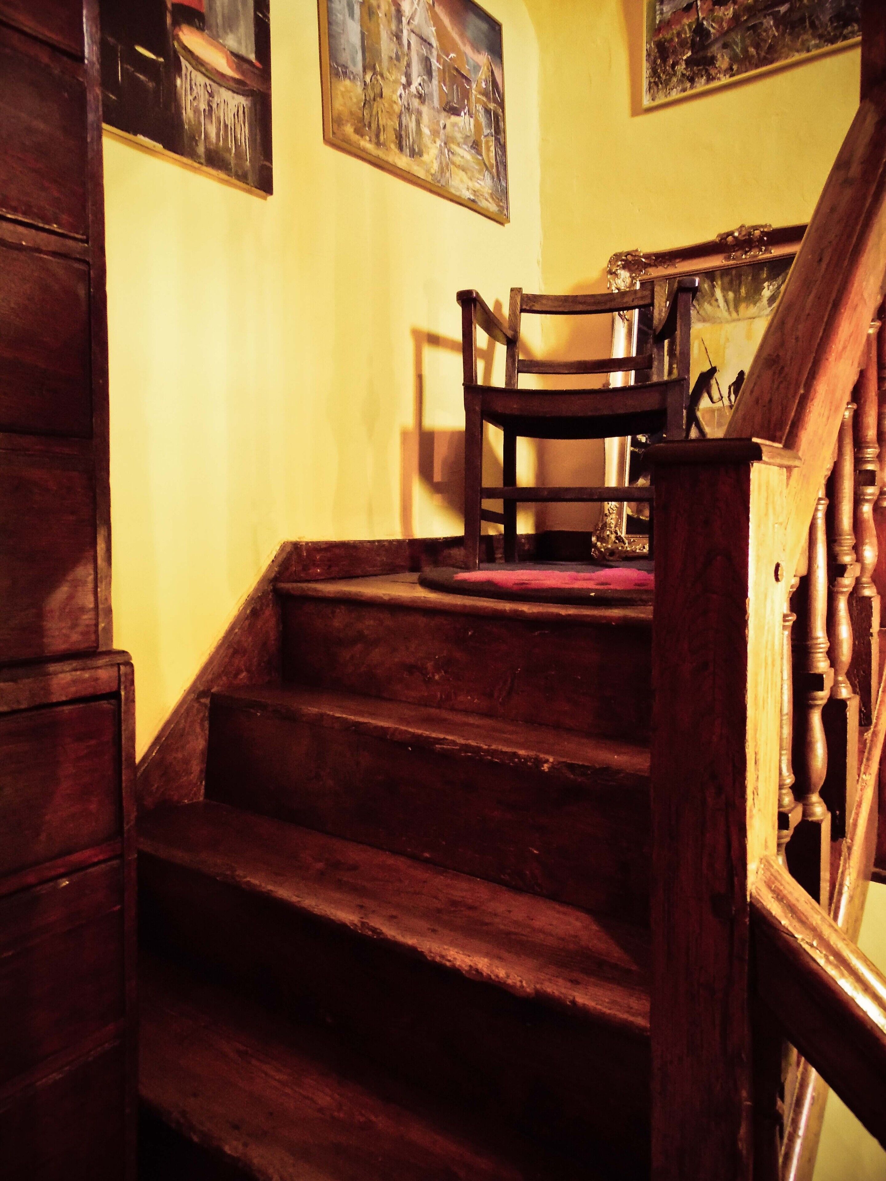 A wooden staircase with a chair at the top, wooden railings, and paintings on the walls.
