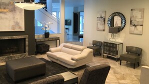 Un soggiorno moderno con camino, divano, poggiapiedi e poltrona con motivo decorativo. Si intravede una scala sullo sfondo.