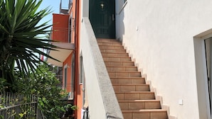 Un escalier menant à l’entrée d’un immeuble, avec une porte verte, le tout entouré de plantes et d’un petit jardin.