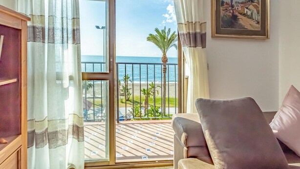 Een kamer met uitzicht op zee, een balkon met een palmboom en een lijst met een schilderij aan de muur.