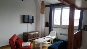 Una sala de estar con un sofá rojo, un sofá azul, una mesa blanca, un baúl antiguo, un televisor y una ventana con cortinas.