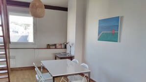 Una habitación con una mesa y sillas blancas, una estantería, un cuadro y una ventana con vistas.