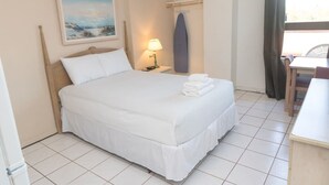 Uma cama bem arrumada com lençóis brancos, uma mesa de cabeceira com abajur, um quadro na parede e uma prancha de surf pendurada.