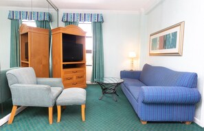 A hotel room with a blue sofa, a grey armchair, a wooden TV stand, a glass coffee table, and a lamp.
