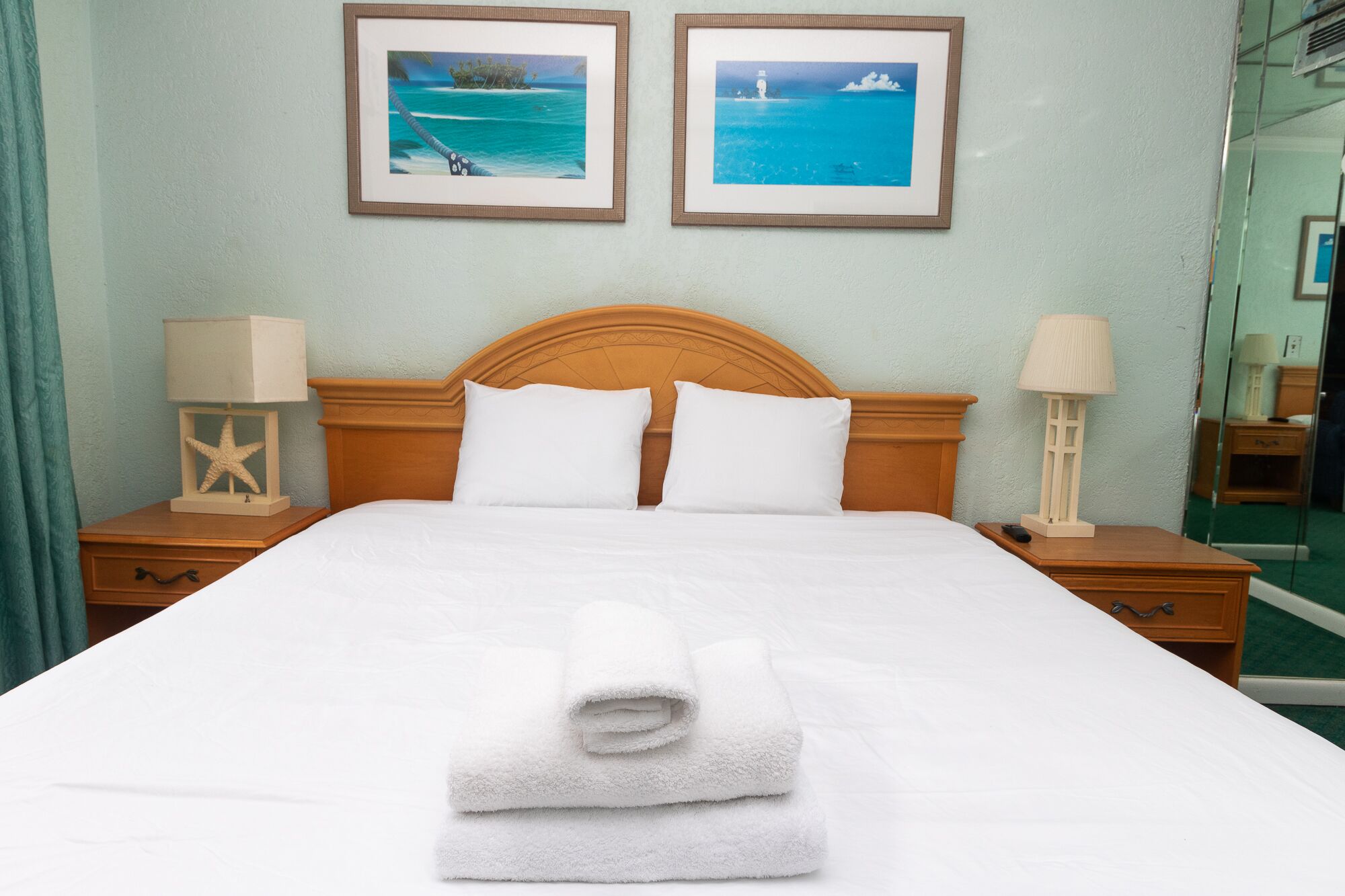 A neatly made bed with white linens and folded towels, a wooden headboard, two bedside tables with lamps, and two framed pictures of tropical scenes on the wall.