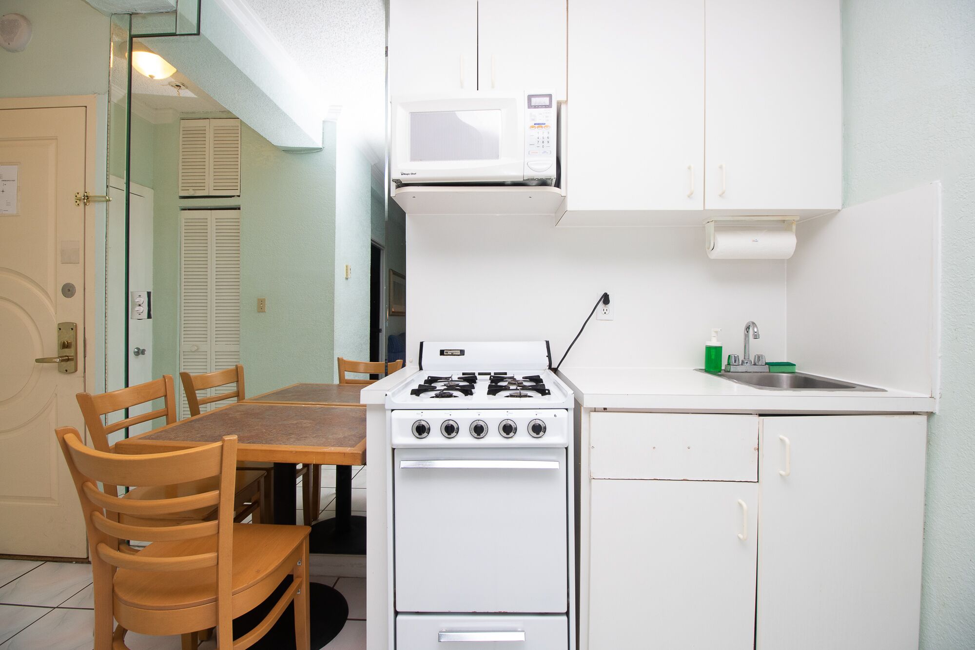 Een compacte keuken met een gasfornuis, een magnetron, een spoelbak en een klein aanrecht. Er staat een houten eettafel met stoelen.