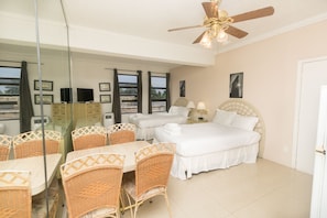 Une chambre d’hôtel avec deux lits, un coin repas avec des chaises en osier, un ventilateur de plafond et une grande fenêtre donnant sur un paysage agréable.