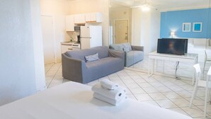 Une chambre d’hôtel moderne avec un canapé gris, un petit coin cuisine, une télévision et un lit avec du linge de maison blanc.