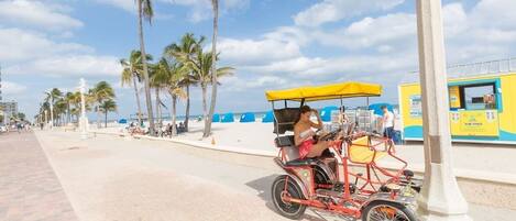 A red three-wheeled vehicle with a yellow canopy, parked on a paved walkway with palm trees and a beach in the background.