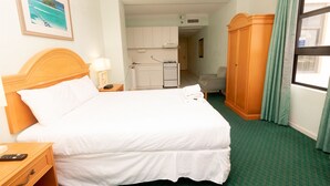 Une chambre d’hôtel comprenant un lit, une table de chevet, une lampe, un tableau, un coin cuisine, une armoire et une fenêtre avec des rideaux.