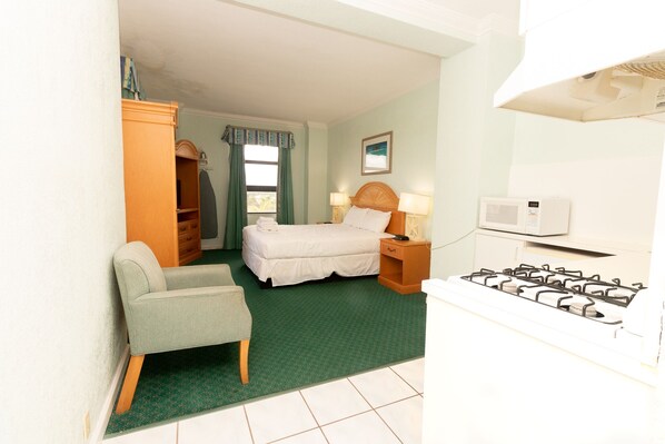 Habitación de hotel con una cama, una silla, un armario, un escritorio con lámpara, microondas y estufa a gas.