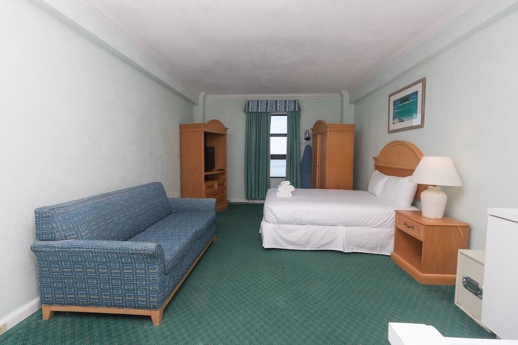 Ein Hotelzimmer mit einem Bett, einem blauen Sofa, einem Holzschrank, einem Nachttisch mit Lampe und einem Fenster mit Vorhängen.