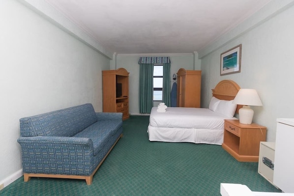 A hotel room with a bed, a blue sofa, a wooden wardrobe, a nightstand with a lamp, and a window with curtains.