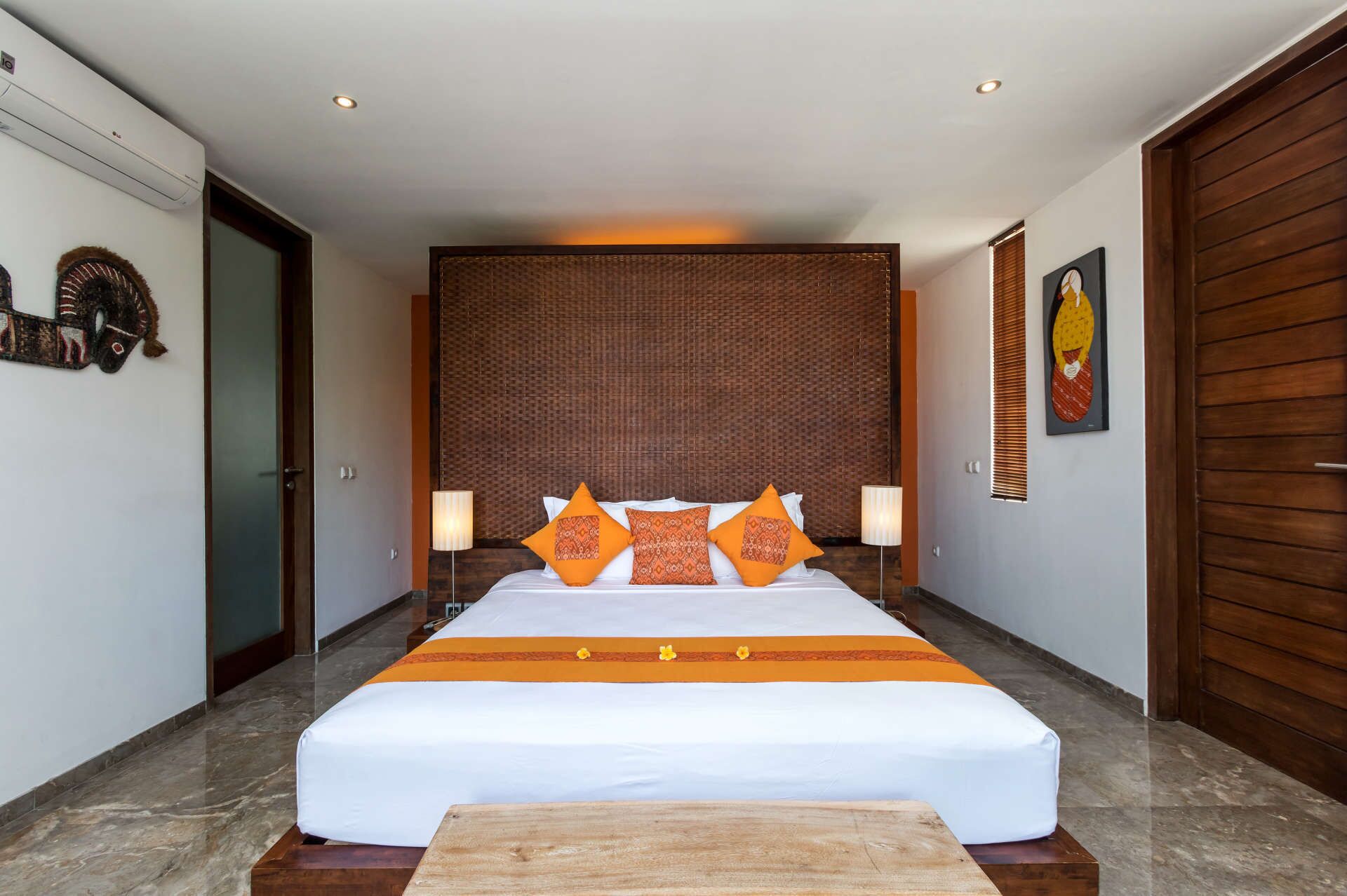 A large bed with white and orange bedding, two bedside lamps, a wooden headboard, and a wooden door on the right.