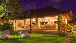 A covered outdoor seating area with two lounge chairs, a dining area with tables and chairs, and a garden with a pathway. - 5 Star Villa for Rent in Bali, Bali Villa 2047 (Bali)