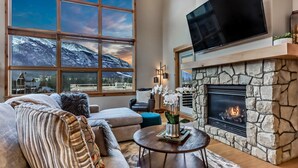 A cozy living room with a stone fireplace, a large window with a mountain view, and a flat-screen TV mounted above the fireplace. - Luxury 3BR/2BA Corner Penthouse in Heart of Canmore (Canmore)