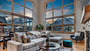 A spacious living room with a large window offering a mountain view, a cozy sofa, a coffee table, and a stone fireplace. - Luxury 3BR/2BA Corner Penthouse in Heart of Canmore (Canmore)