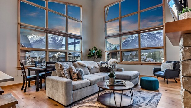 A spacious living room with a large window offering a mountain view, a cozy sofa, a coffee table, and a dining area. - Luxury 3BR/2BA Corner Penthouse in Heart of Canmore (Canmore)