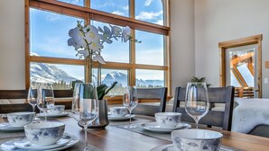 A dining table set with white and blue floral tableware, a vase with white flowers, and a view of snow-capped mountains through large windows. - Luxury 3BR/2BA Corner Penthouse in Heart of Canmore (Canmore)
