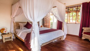 A four-poster canopy bed with a wooden headboard, a nightstand with a decorative item, and a view of greenery through a window with red curtains.