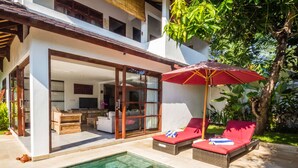 A modern house with a glass door leading to a living area, a pool, and outdoor seating with a red umbrella.