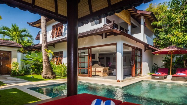 A two-story house with a pool, a covered patio, and a view of the surrounding greenery.