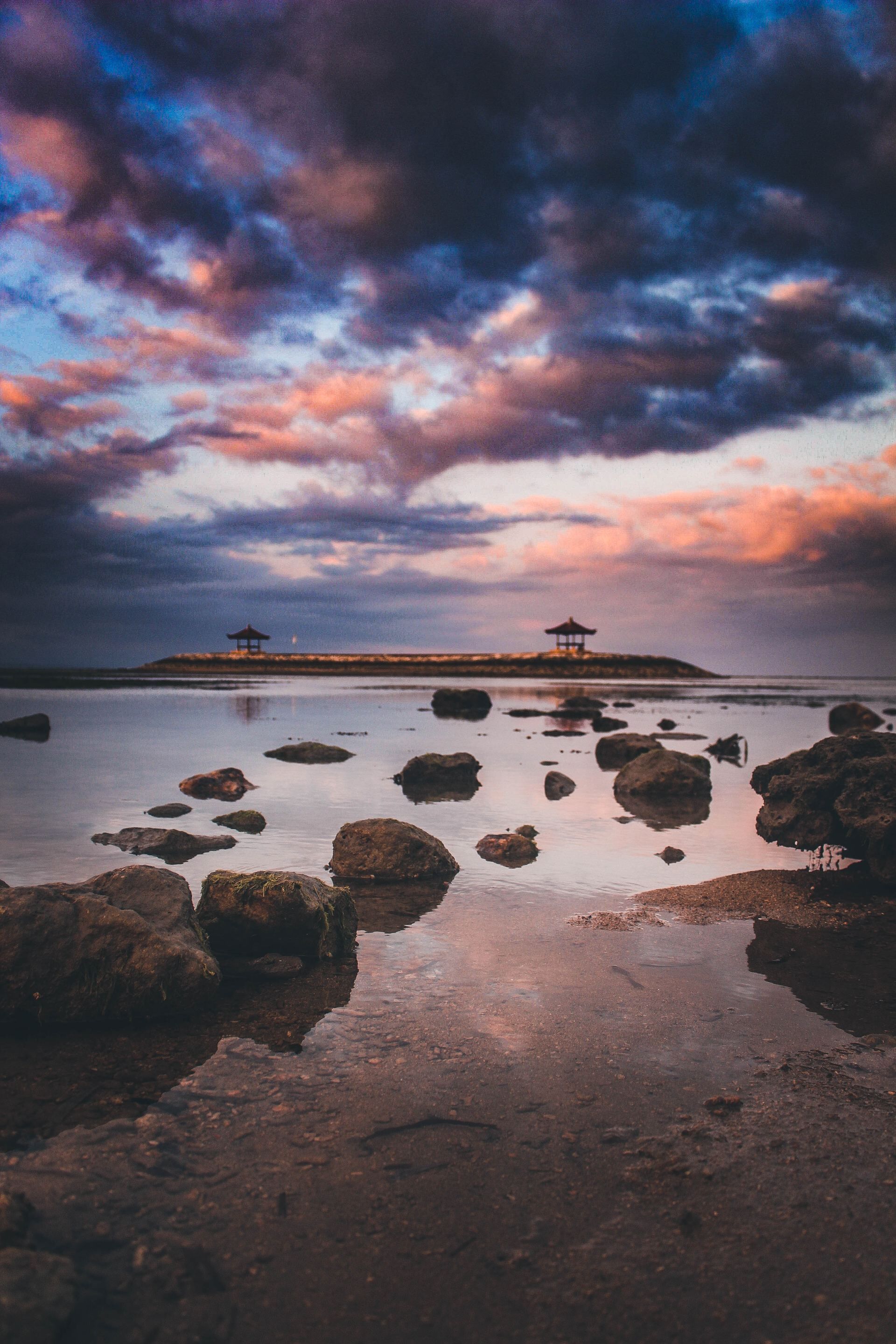 A tranquil beach scene with two gazebos on a small island, surrounded by calm waters and scattered rocks.