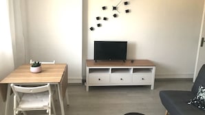 Ein kleiner, ordentlicher Wohnraum mit einem Holzespeisetisch, einem weißen Stuhl, einer Couch, einem Fernseher auf einem Holzboden und einer Wanduhr.