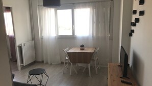 Ein Zimmer mit einem Holzesstisch, weißen Stühlen, einem kleinen runden Tisch, einem schwarzen Stuhl, einem Schreibtisch aus Holz mit Fernseher und Fernbedienung, einem Heizkörper und einem Fenster mit durchsichtigen Vorhängen.